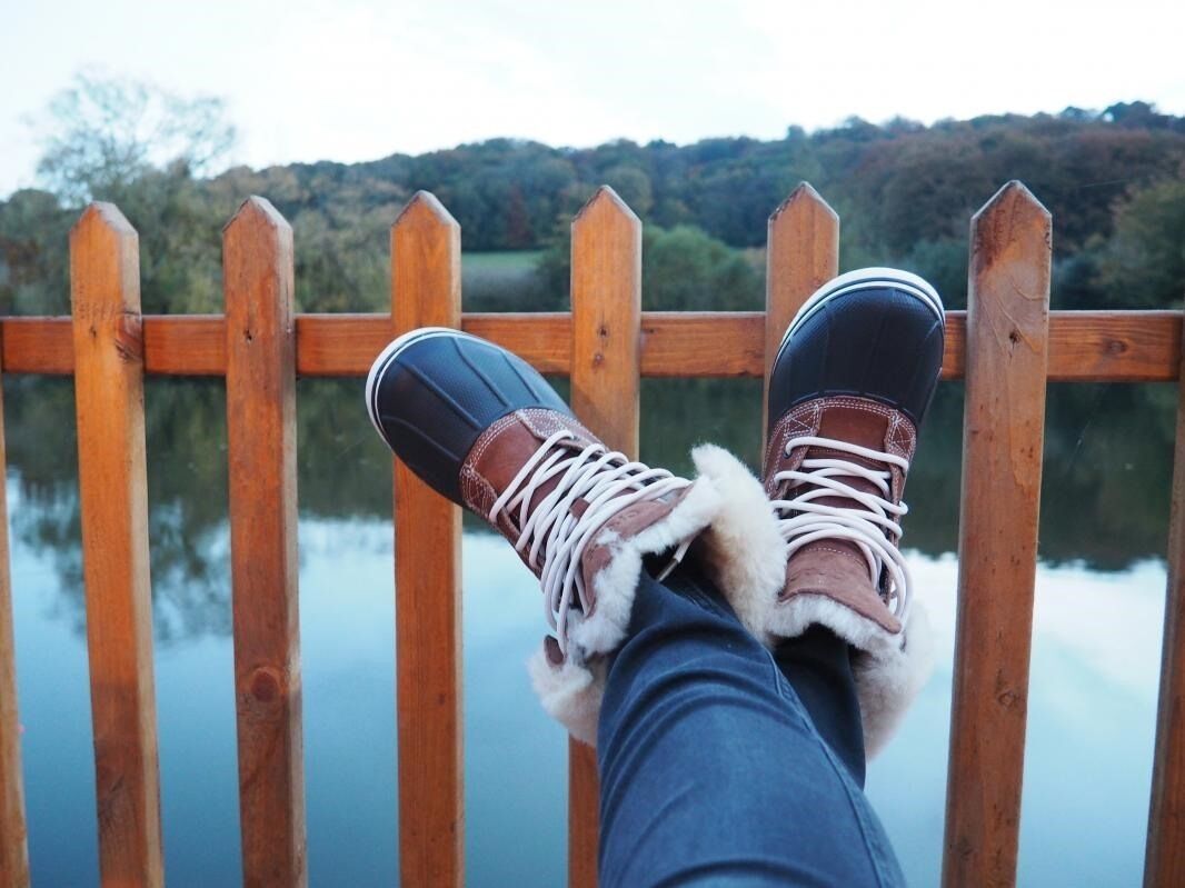 A wooden deck railing with a person resting their feet on it, overlooking a calm body of water and a forested hillside.