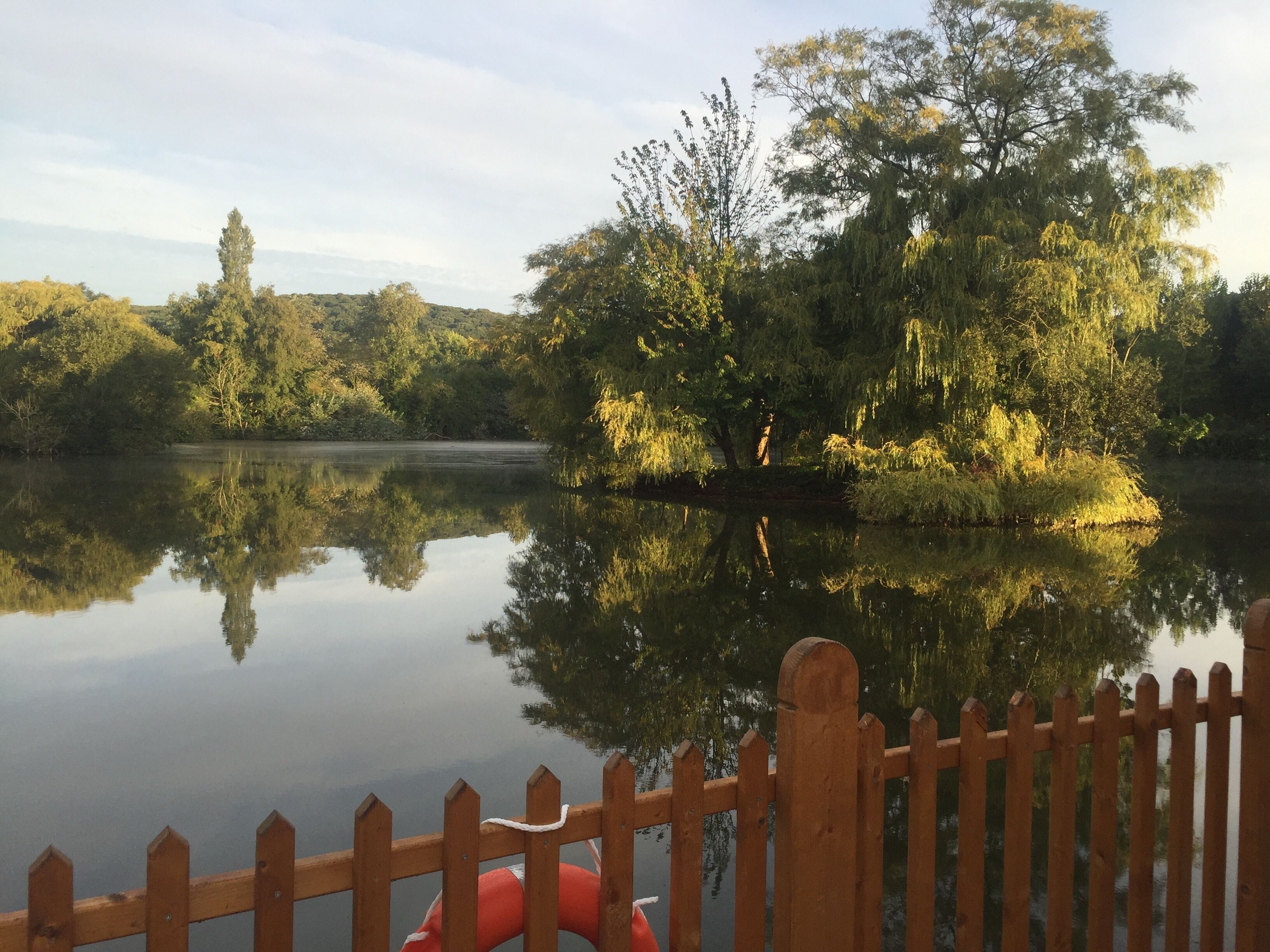 A calm lake with a wooden fence in the foreground and trees reflecting on the water.