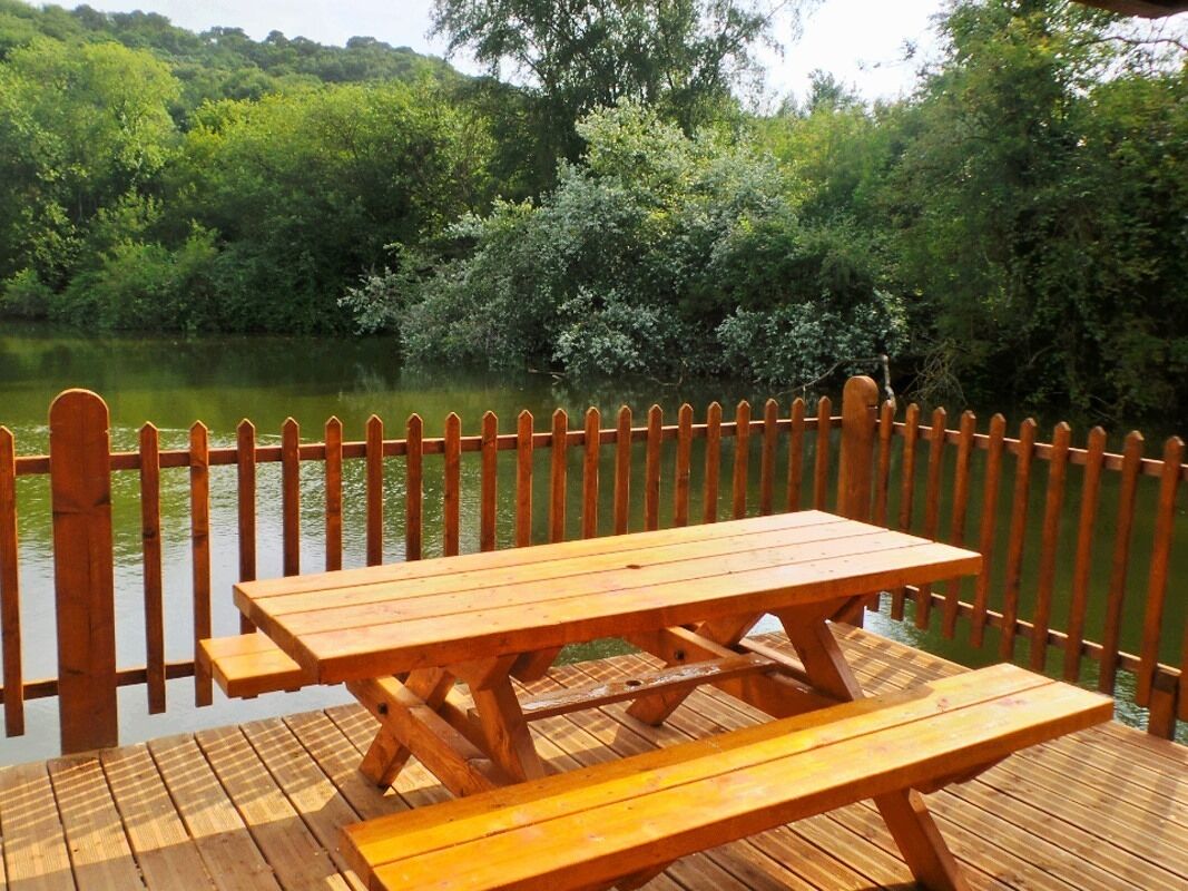 Une terrasse en bois avec des tables de pique-nique, donnant sur un lac et entourée d’une clôture en bois.