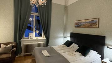 A hotel room with a large bed, a chair, a radiator, a window with curtains, and a chandelier.