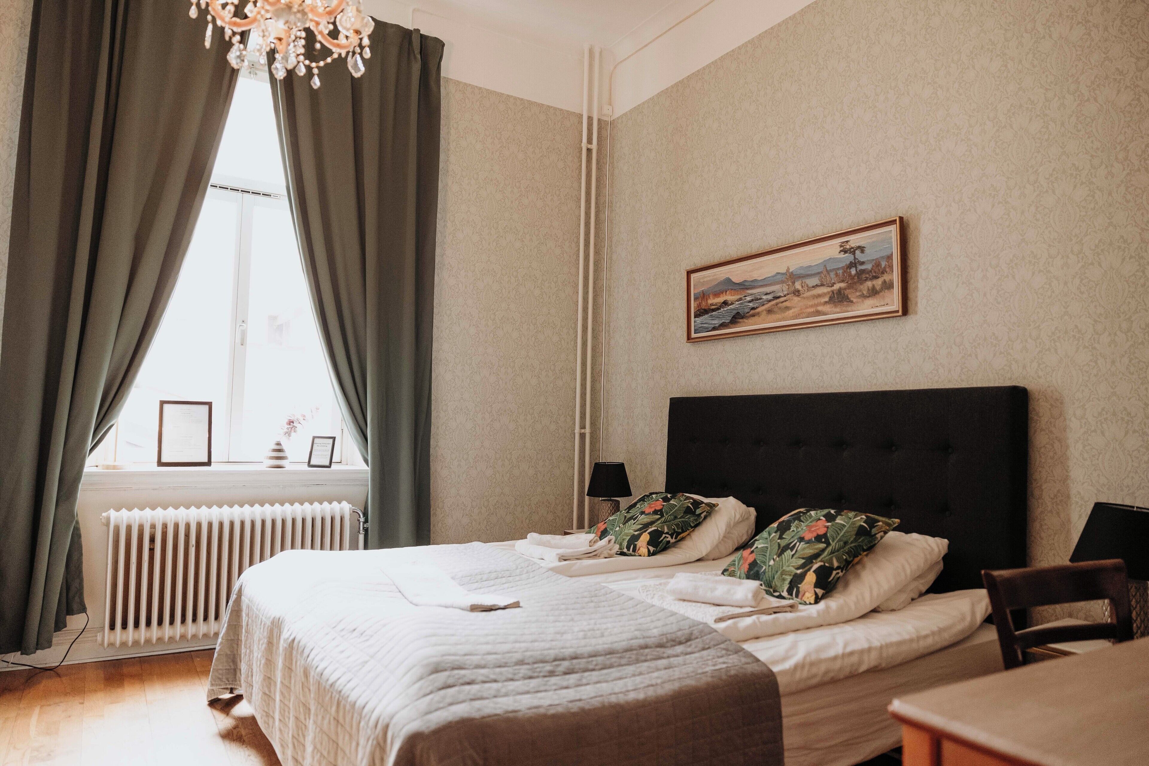 A neatly made bed with pillows, a radiator, a window with curtains, a painting on the wall, and a chair.