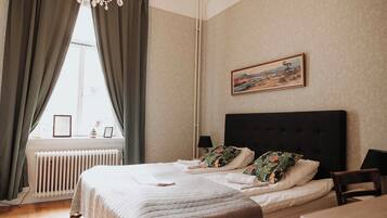 A neatly made bed with pillows, a radiator, a window with curtains, a painting on the wall, and a chair.