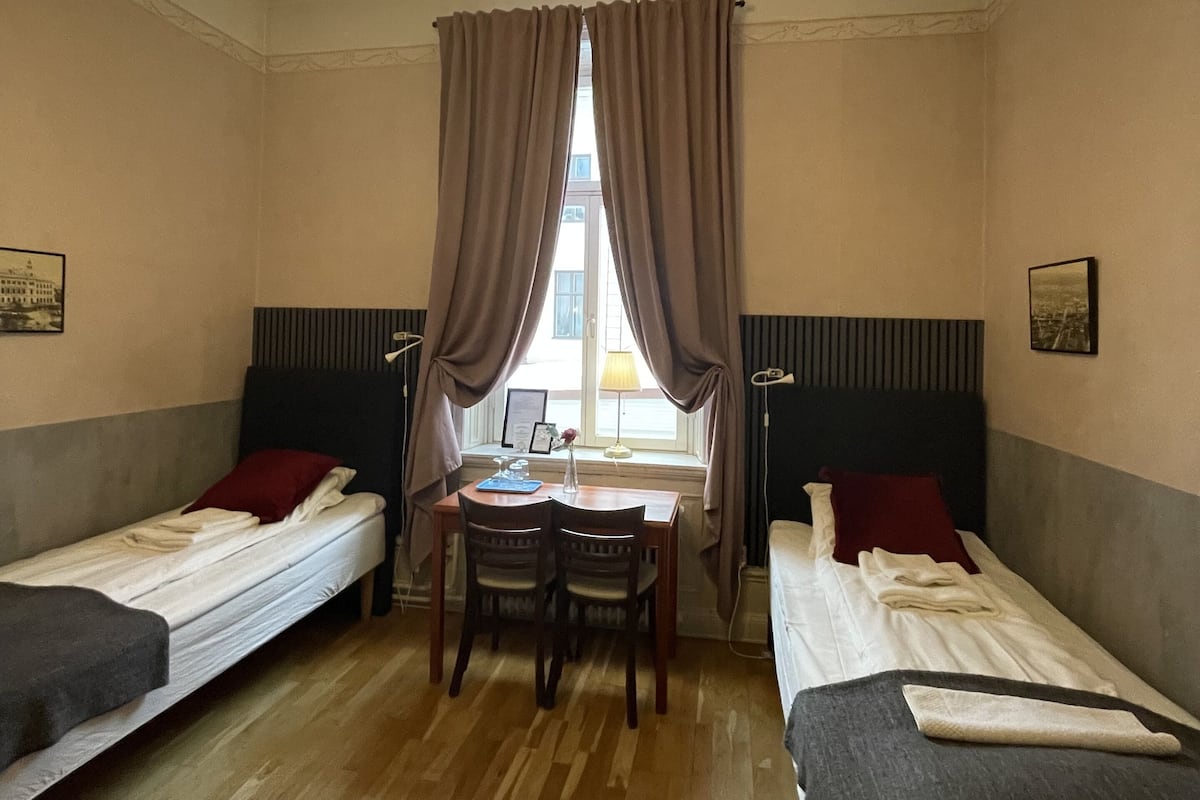 Una habitación con dos camas individuales, una mesita con sillas, un ventanal con cortinas y una lámpara colgante.