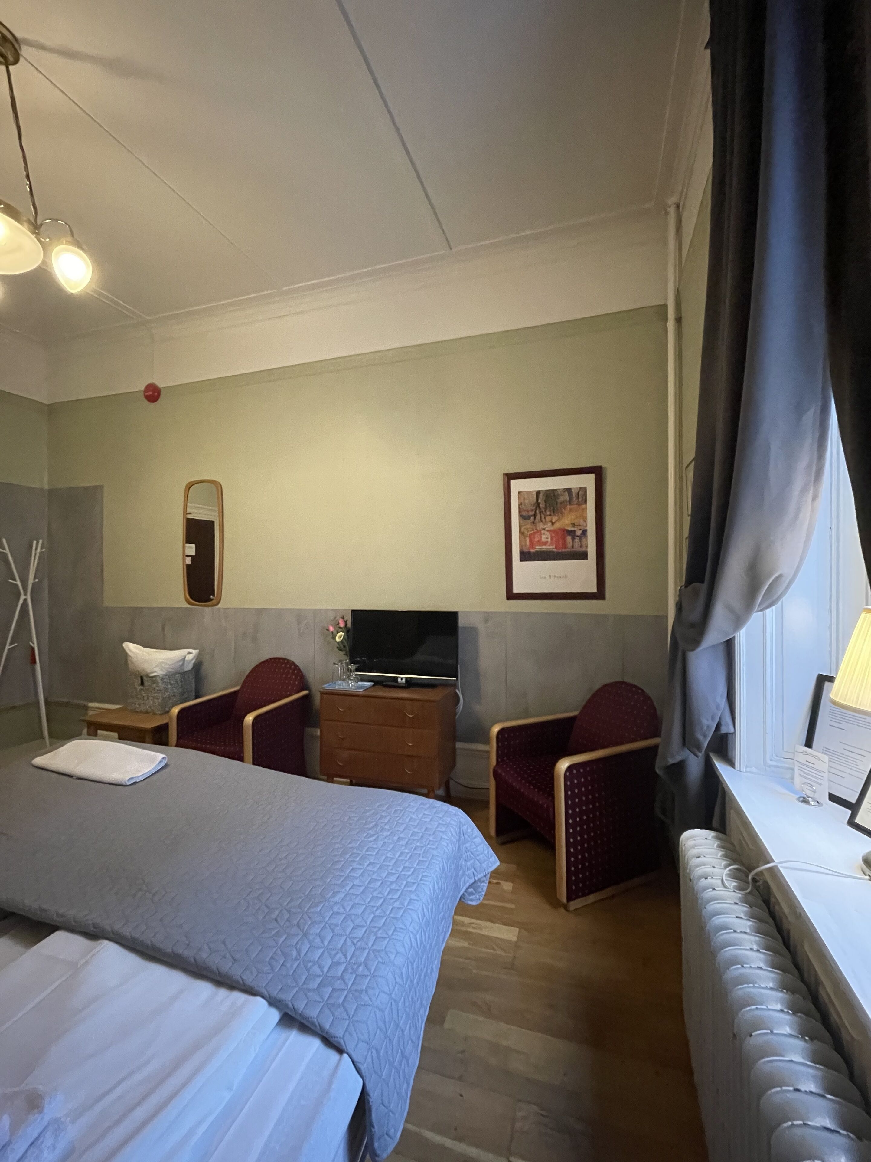 Ein Hotelzimmer mit einem Bett, zwei Sesseln, einer Holzkommode, einem Fernseher und einem Fenster mit Vorhängen.