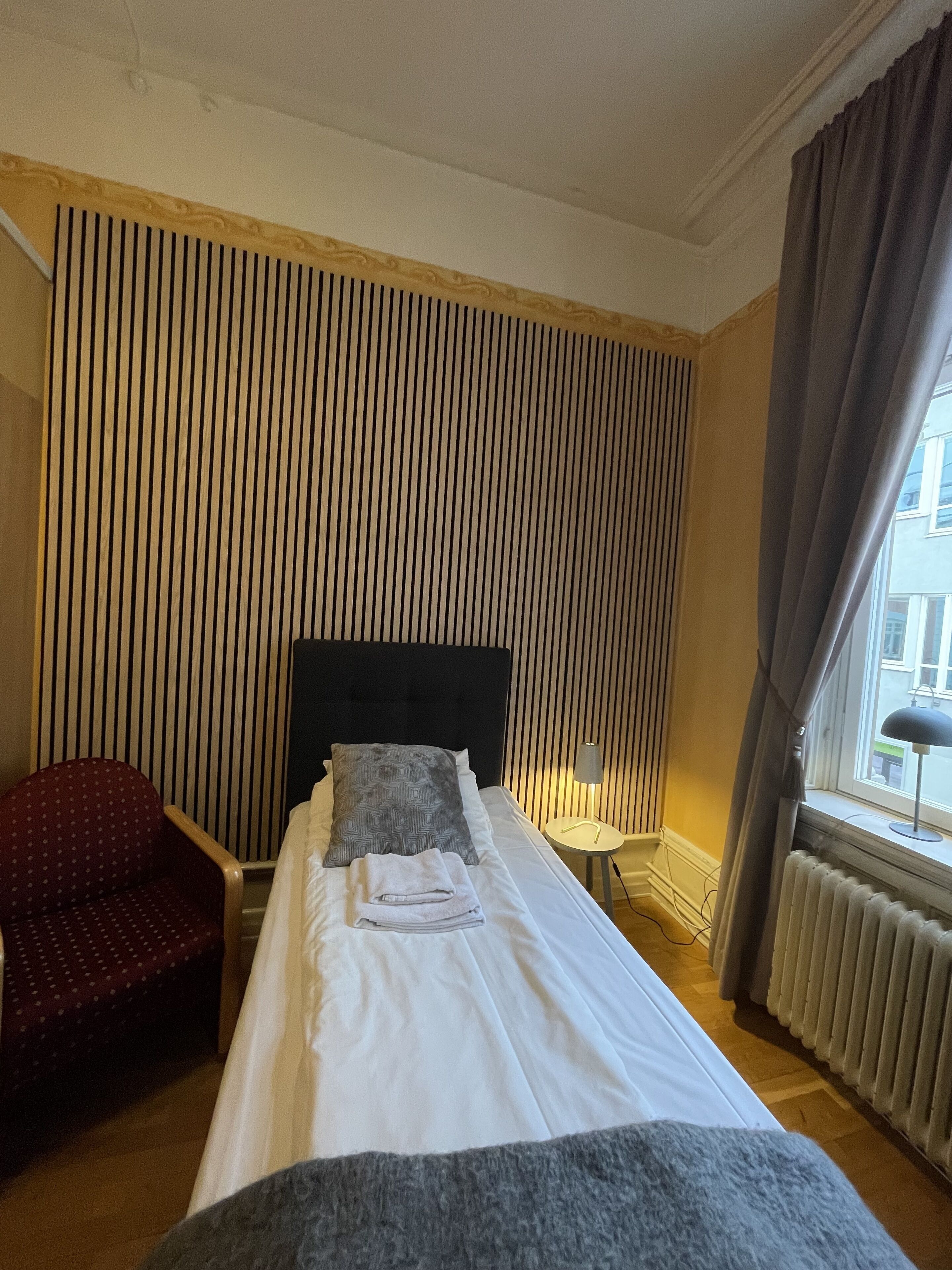 Une chambre d’hôtel avec un lit, une chaise, une lampe et une fenêtre avec des rideaux.
