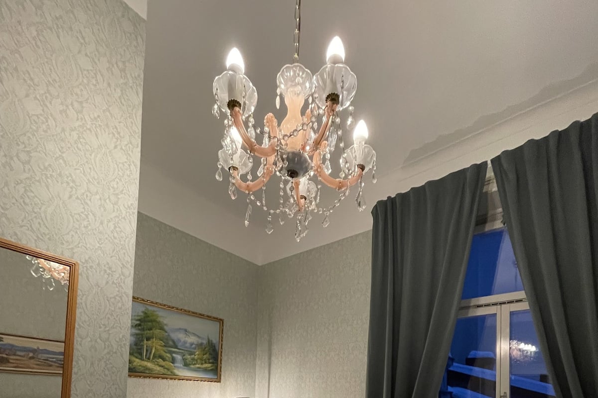 Habitación de hotel con candelabro, dos sillones, un sofá, un cuadro en la pared, una ventana con cortinas y un radiador.