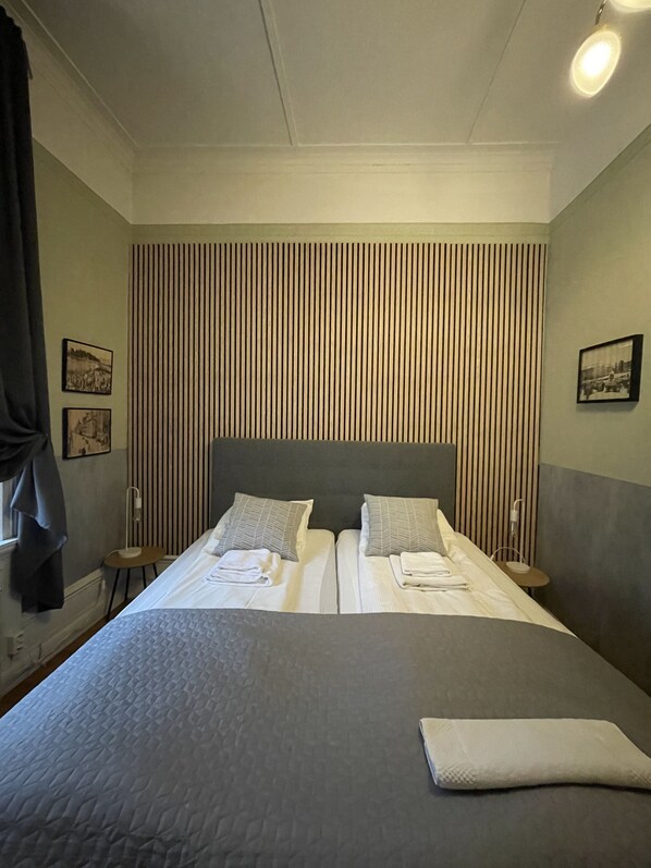 Una cama doble con cabecera gris, dos almohadas y una toalla doblada a cada lado. Hay dos mesitas de noche con lámparas. La habitación tiene un panel de pared a rayas y dos cuadros enmarcados.