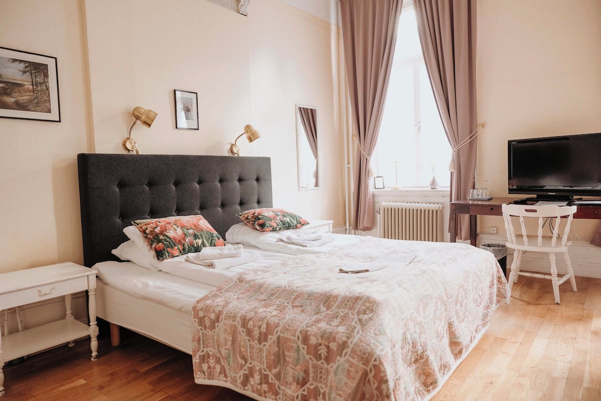 Un dormitorio ordenado con cama, mesitas de noche, un escritorio con silla, televisión y ventana con cortinas.
