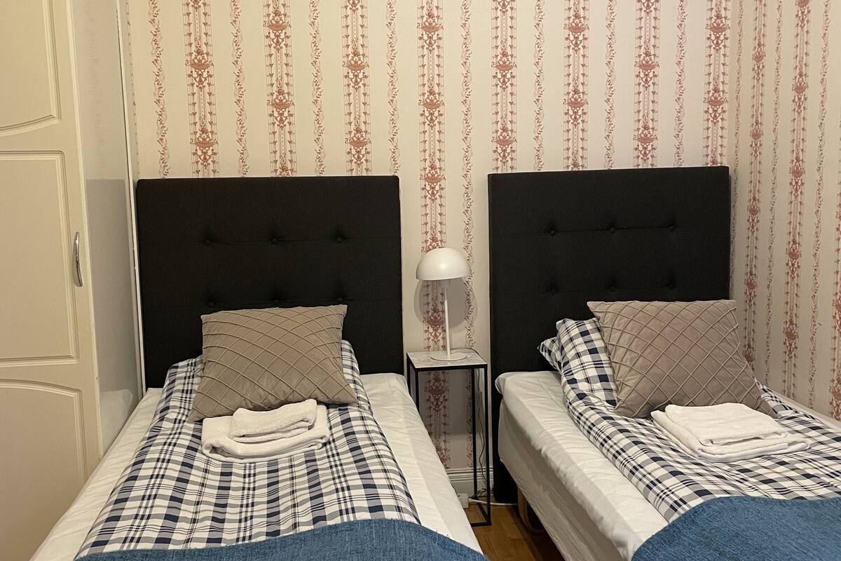 Habitación de hotel con dos camas, cada una con cabecera, mesita de noche y lámpara. La habitación tiene papel tapiz estampado y una puerta a la izquierda.