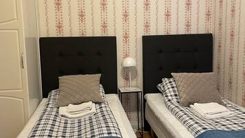 A hotel room with two beds, each with a headboard, a nightstand, and a lamp. The room has patterned wallpaper and a door on the left.