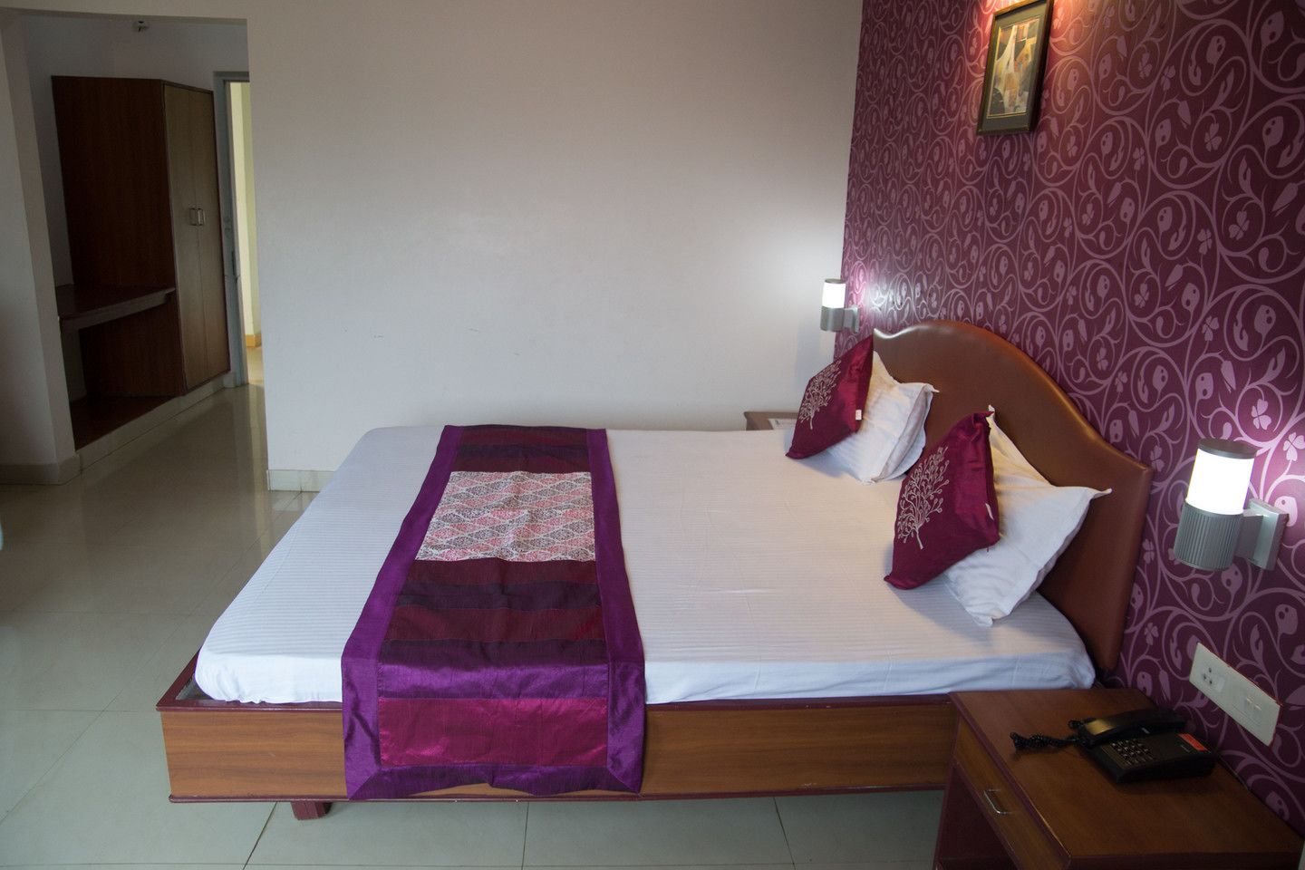 A neatly made bed with a purple blanket and pillows, a bedside table with a lamp, and a patterned wall.