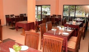 A restaurant with multiple tables and chairs, large windows, and a view of greenery outside. - Hotel New Rockbay (Puri)