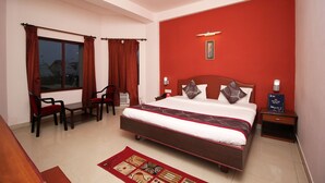 Habitación de hotel con una cama grande, un escritorio, dos sillas, una pared roja, un ventilador de techo y una ventana con cortinas.