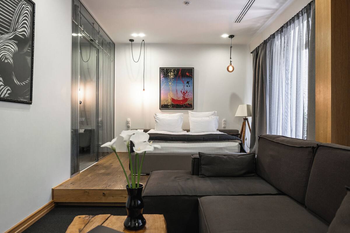 Una habitación de hotel moderna con una cama, un sofá, una mesita de madera, un jarrón con flores y un cuadro en la pared.