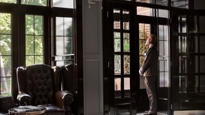 A man standing at a black door with glass panels, a leather armchair, and a small round table with a book. - Wall Street - Maestro (Odesa)