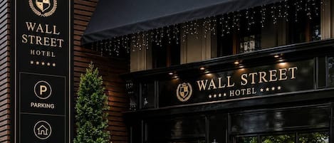 Exterior del Hotel Wall Street, con fachada negra, un cartel que enumera servicios como estacionamiento, refugio, política de mascotas y generador eléctrico, y una escalera que conduce a la entrada.