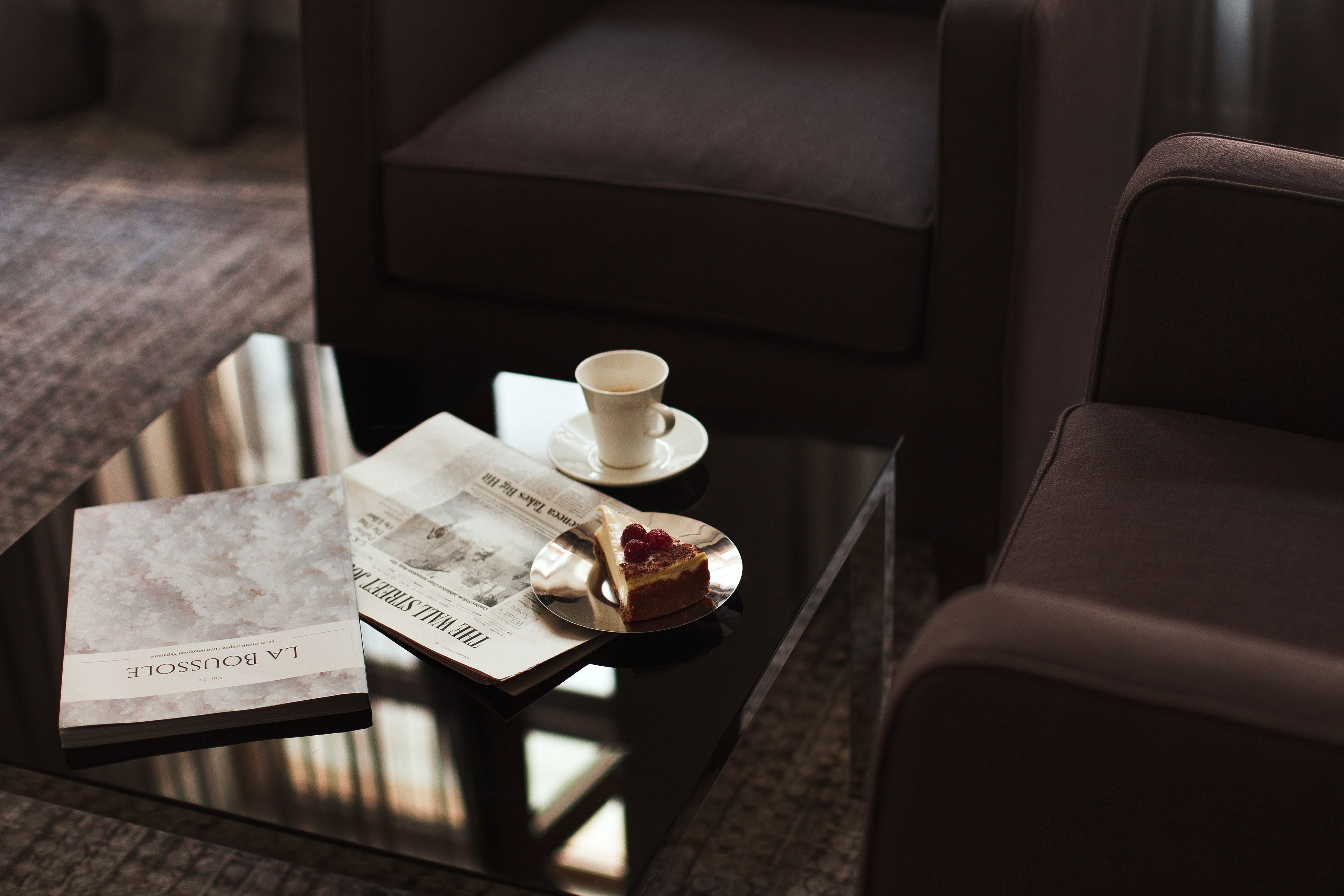 Habitación de hotel con una mesa de vidrio que sostiene un libro, una taza y una rebanada de pastel.