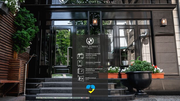 The exterior of the Wall Street Hotel with a clear entrance, steps leading up to the door, and a signboard with icons indicating amenities such as a generator, autonomous heating, water supply, and shelter. - Wall Street - Maestro (Odesa)