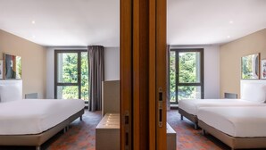 Habitación de hotel con dos camas, una mesa central y amplios ventanales con vistas a zonas verdes.