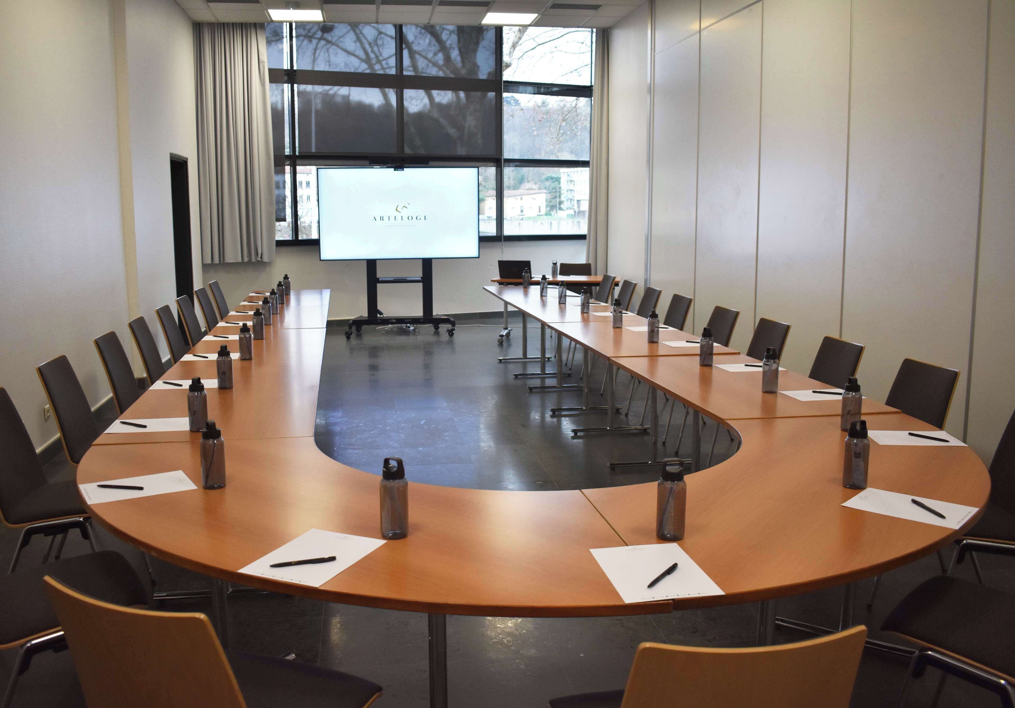 Ein Konferenzraum mit einem langen Tisch, Stühlen, Wasserflaschen und einer Präsentationsleinwand.