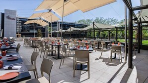 Un área de comedor al aire libre con mesas y sillas, grandes sombrillas y un edificio al fondo.