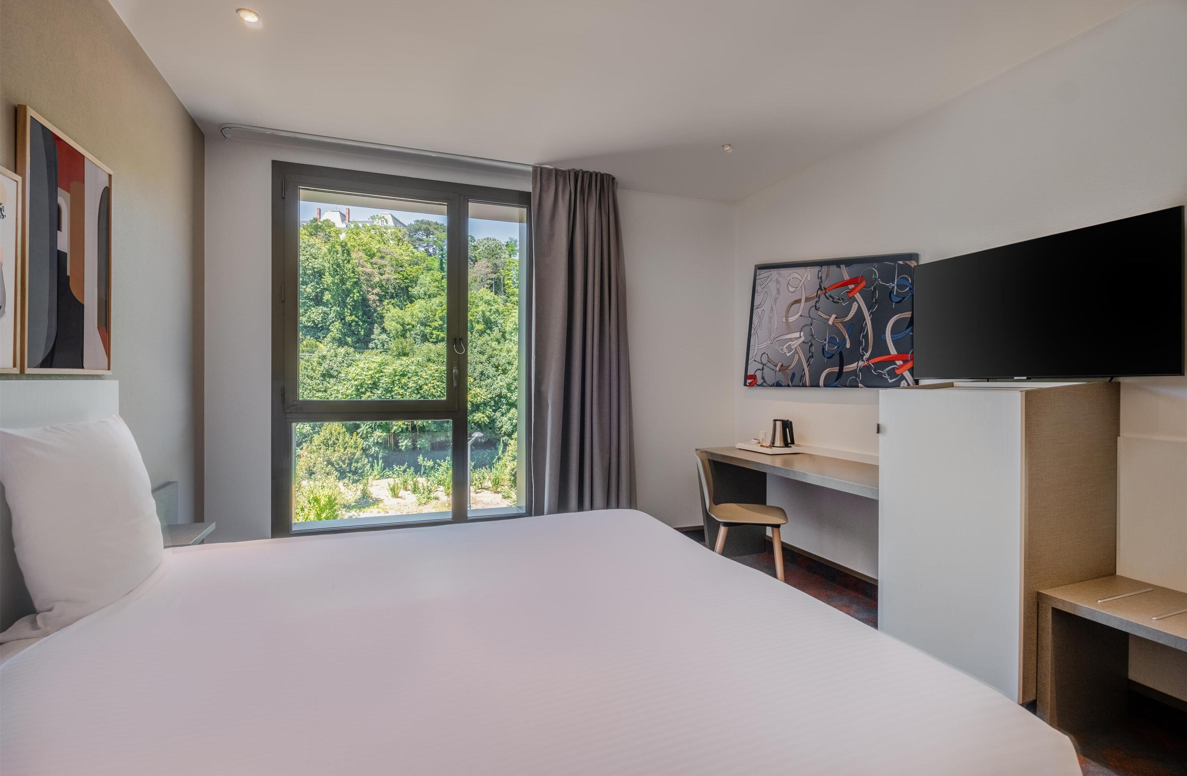 Een moderne hotelkamer met een groot bed, een bureau, een stoel, een flatscreen televisie en een raam met uitzicht op groen.