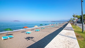 A beach with rows of sun loungers and umbrellas, a concrete walkway, and a clear view of the ocean and distant mountains. - Bahar Hotel (+12 Adult ) (Fethiye)