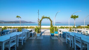 An outdoor seating area with white tables and chairs, a central archway with greenery, and a view of the sea and mountains in the background. - Bahar Hotel (+12 Adult ) (Fethiye)