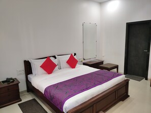 Een keurig opgemaakt bed met een paarse plaid, twee rode kussens en een houten hoofdbord. Er is een nachtkastje met een lamp, een klein bankje en een spiegel aan de muur.
