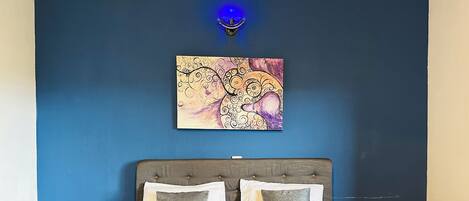 A bedroom with a blue wall, a bed with a grey headboard, and a painting above the bed.