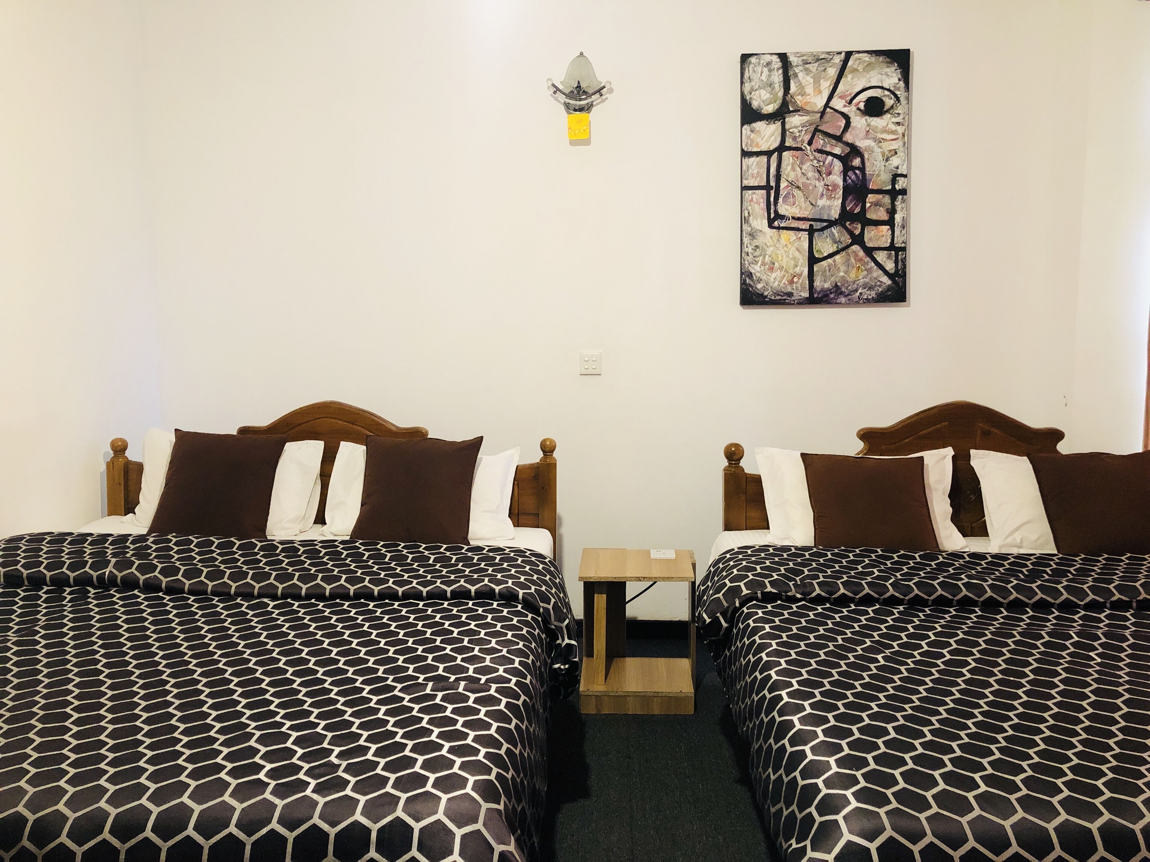 Une chambre avec deux lits, chacun doté d’une tête de lit en bois et d’une couvre-lit à motifs. Une petite table de chevet en bois est placée entre les deux lits, et un luminaire mural est fixé au-dessus des lits.