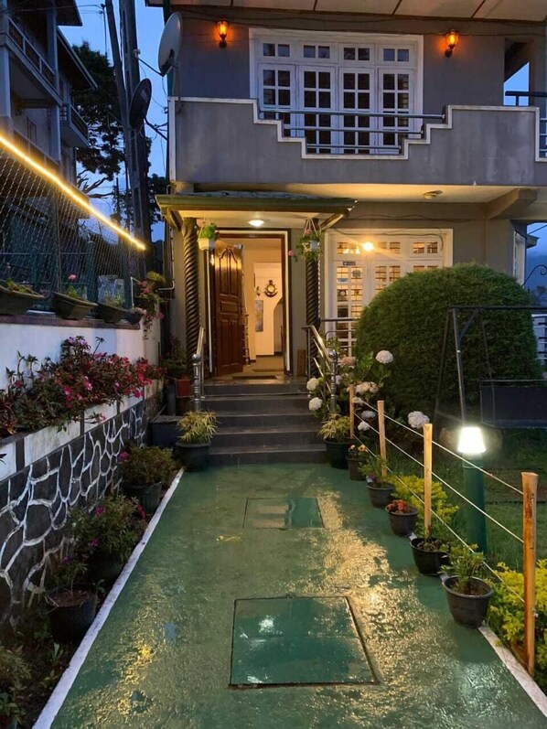 A two-story building with a covered entrance, a staircase leading to the door, and a green pathway with potted plants.
