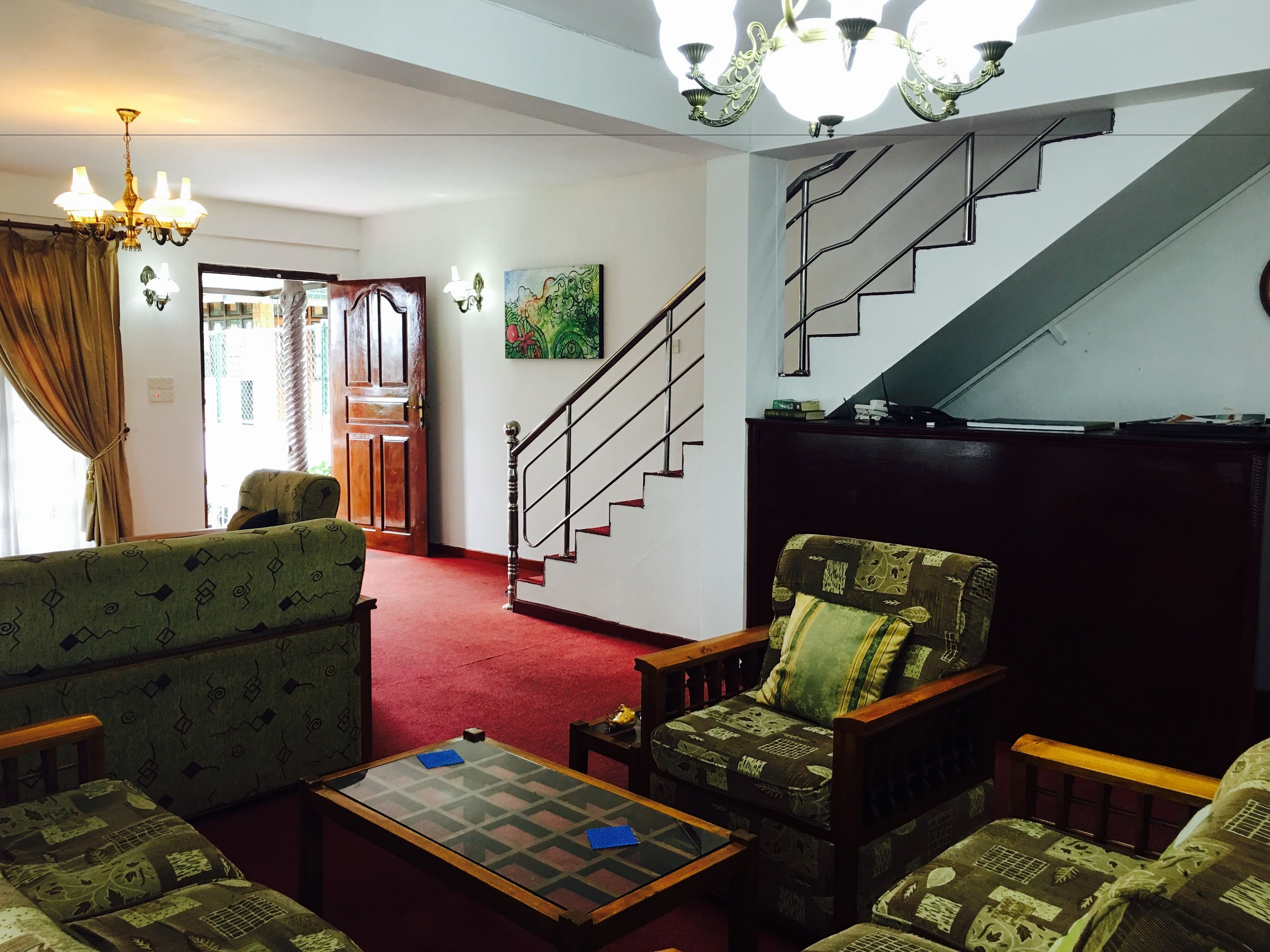 A living room with a red carpet, patterned sofas, a glass-top coffee table, a wooden armchair, and a staircase with a metal railing.