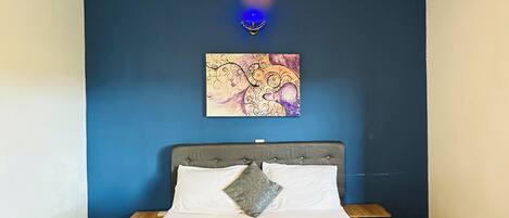 Ein Schlafzimmer mit einem Bett, einem Kopfteil, einem Nachttisch, einer Wandlampe und einem Gemälde.