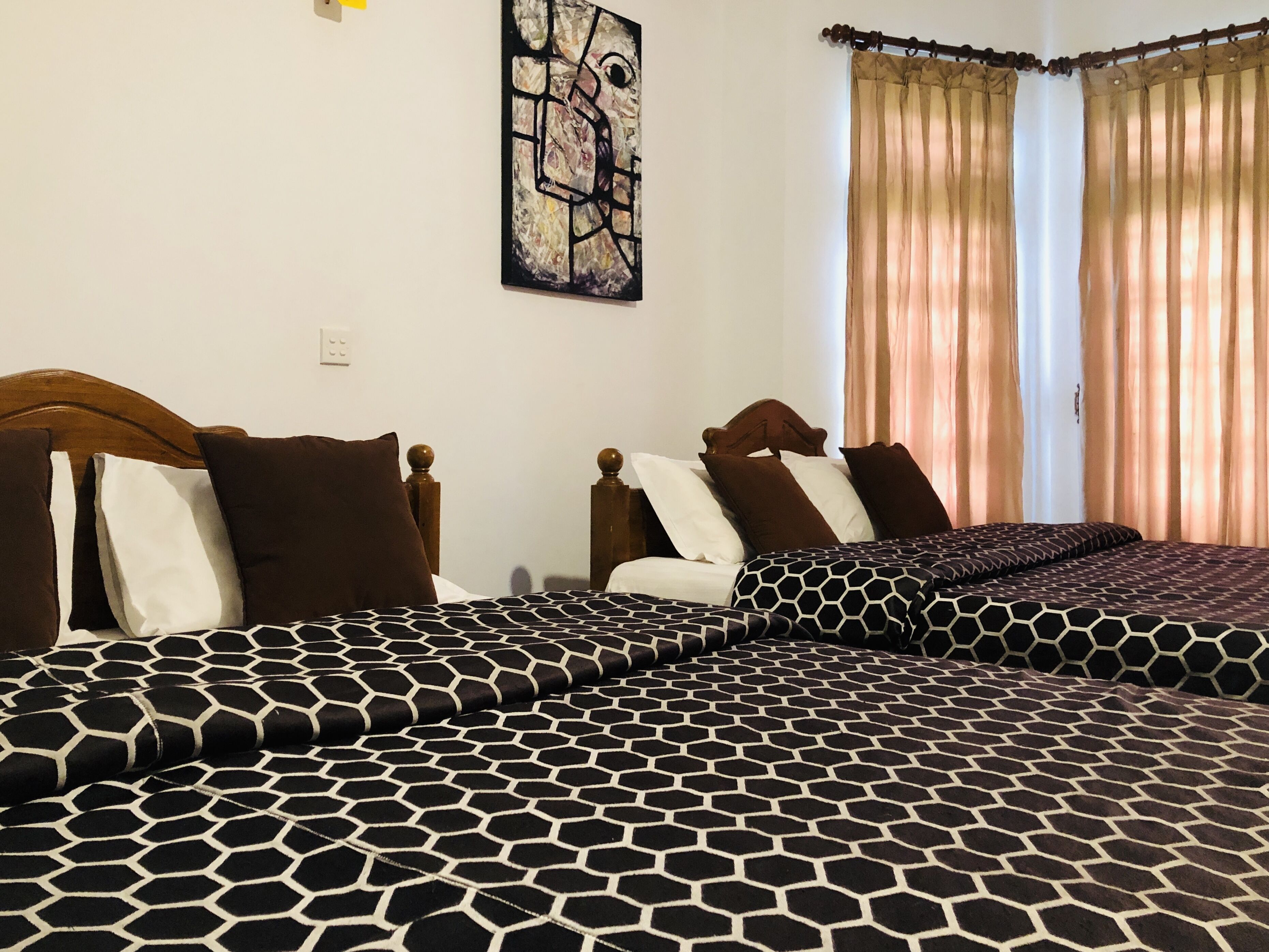 A hotel room with two beds, each with a wooden headboard and a patterned bedspread. There is a painting on the wall and a window with curtains.