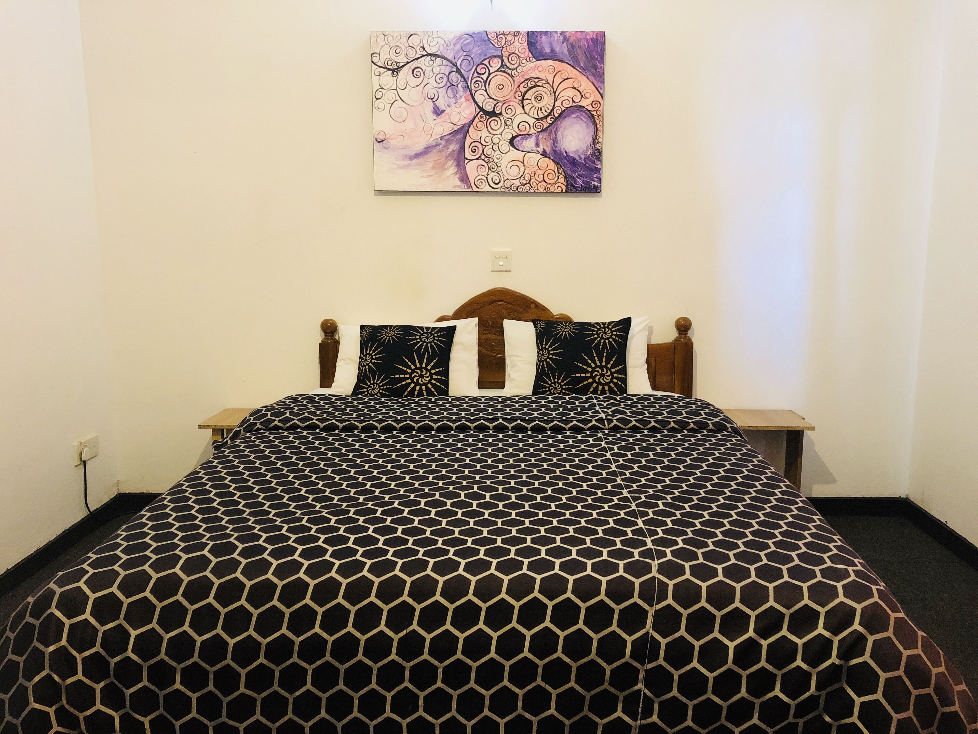 Een net opgemaakt bed met een sprei met een geometrisch patroon, een houten hoofdbord en een abstract schilderij aan de muur.
