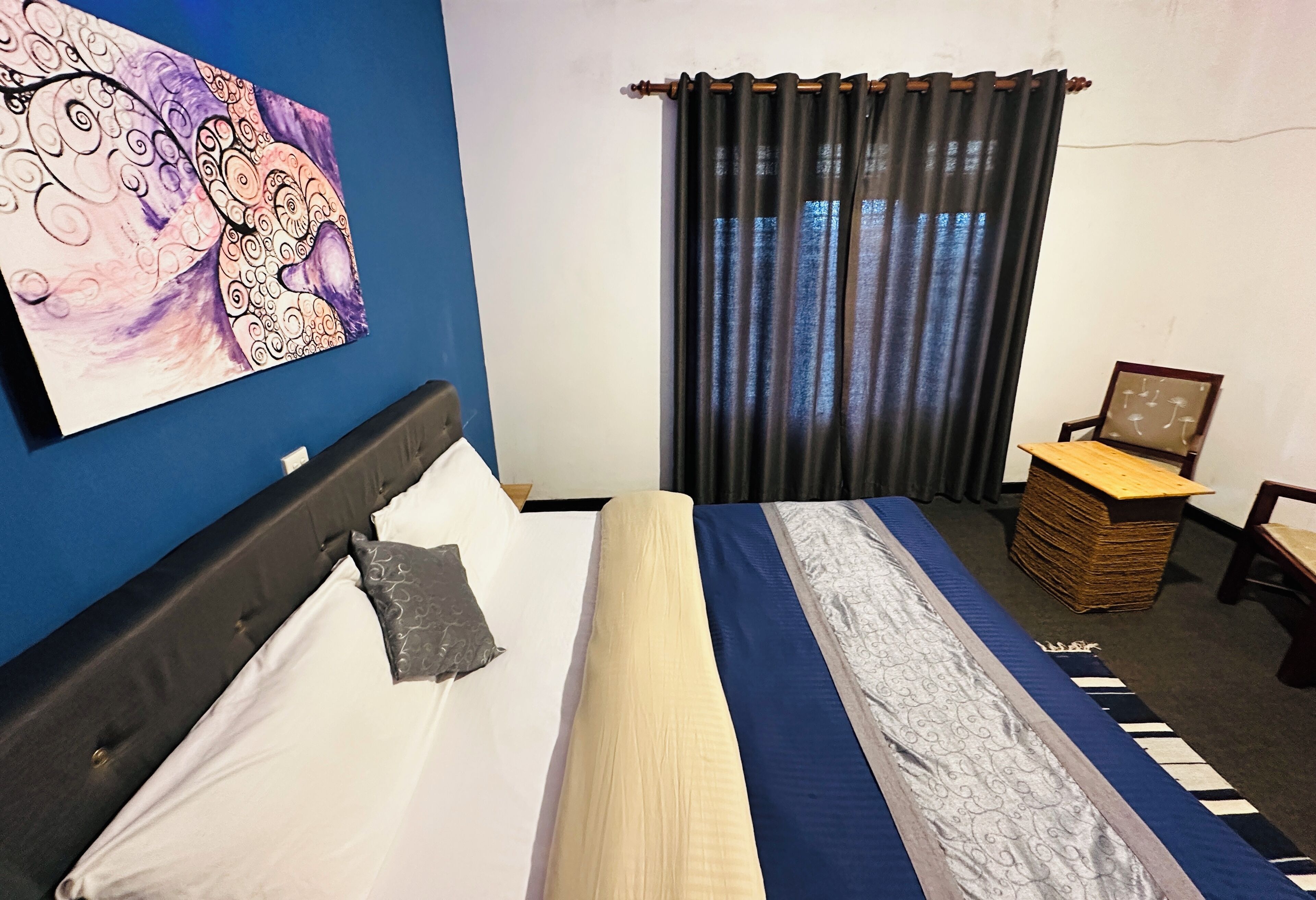 Ein ordentlich gemachtes Bett mit einer blau-weißen, gestreiften Tagesdecke, einem Kissen und einem Kopfteil. An der Wand hängt ein gerahmtes Bild und neben dem Bett steht ein kleiner Holztisch mit einem Stapel Bücher.