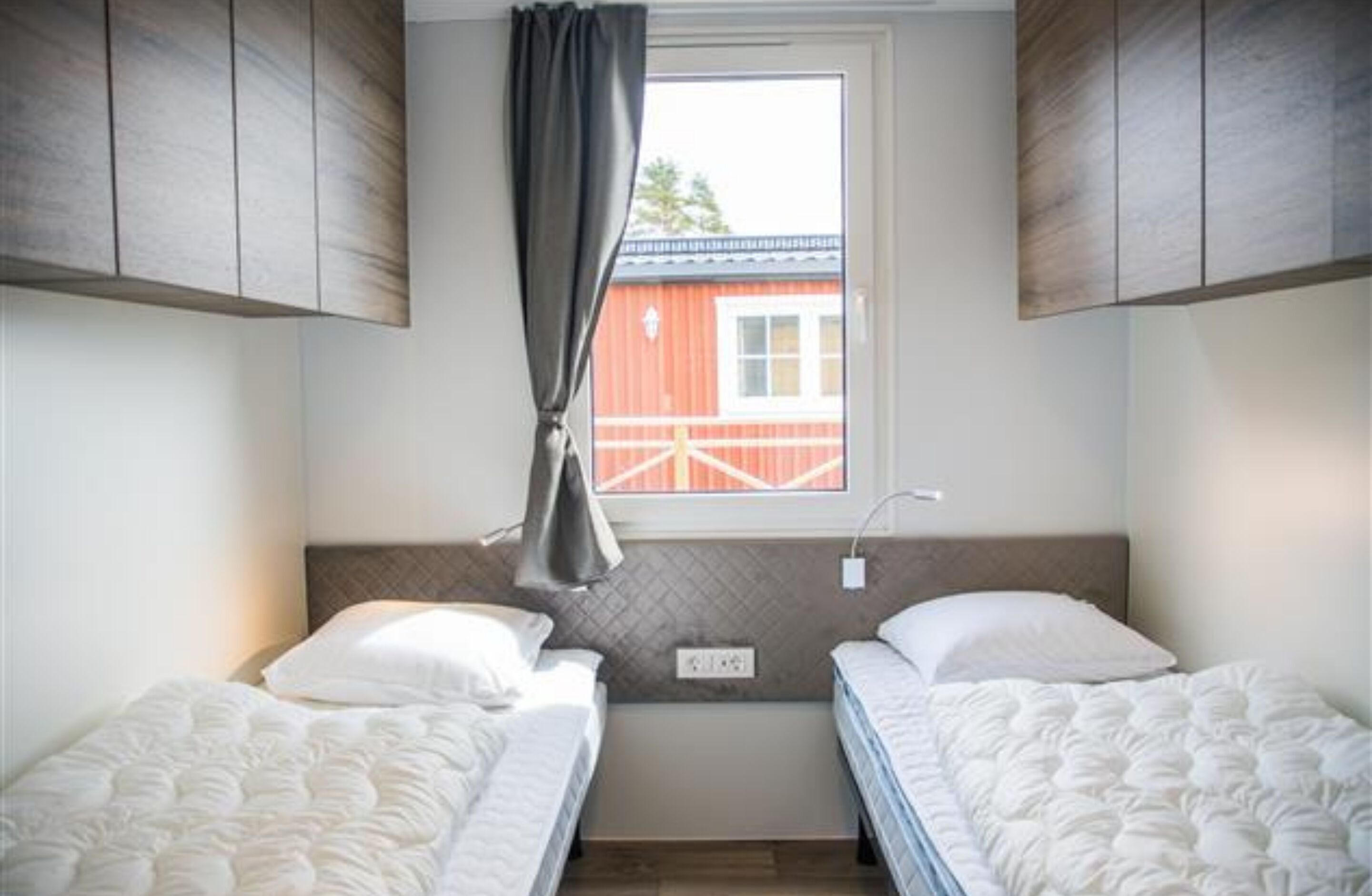A small, well-lit room with two beds, a window with a view of a red building, and a curtain.