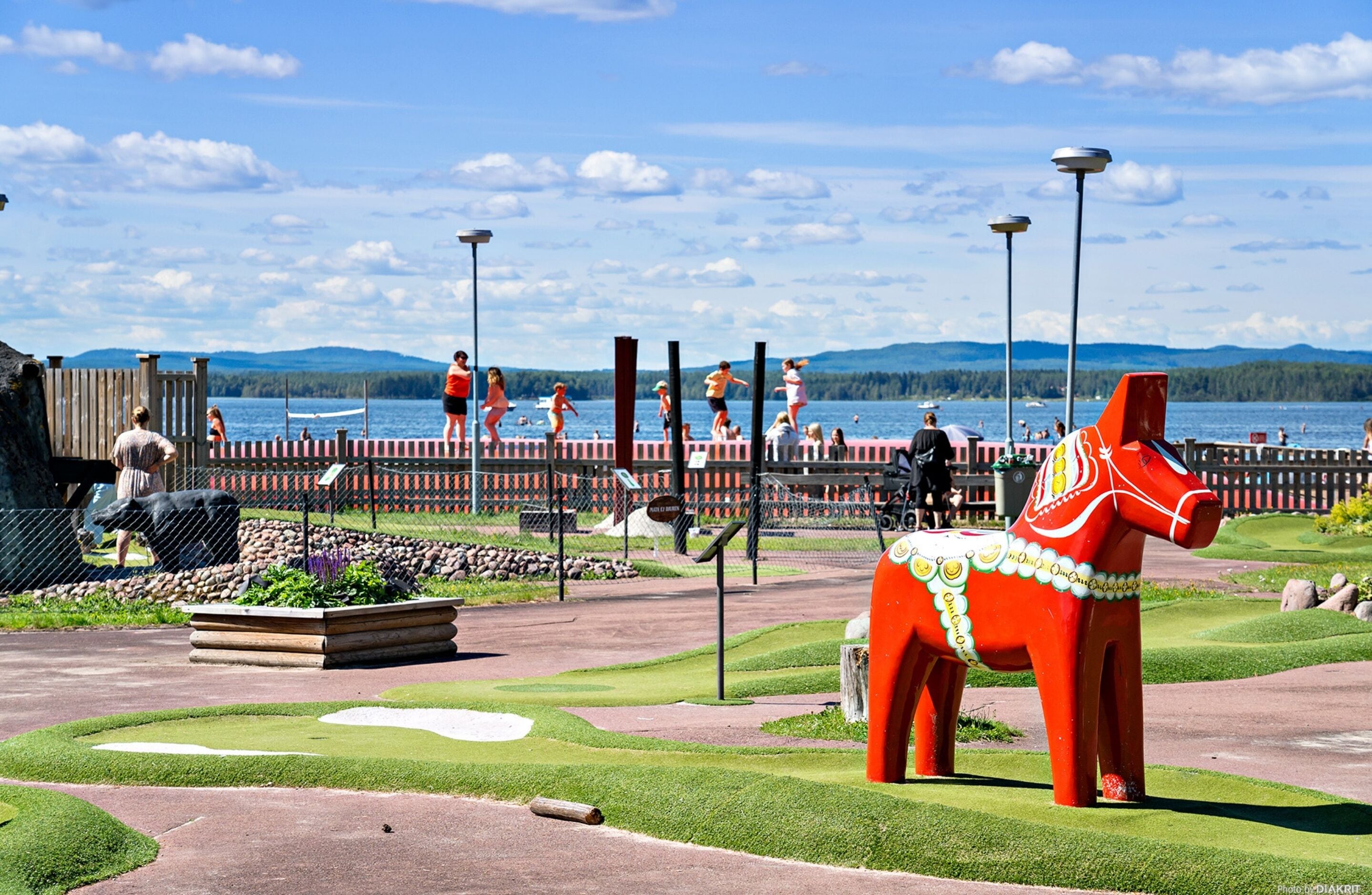 A park with a red horse statue, a wooden fence, and a body of water in the background.