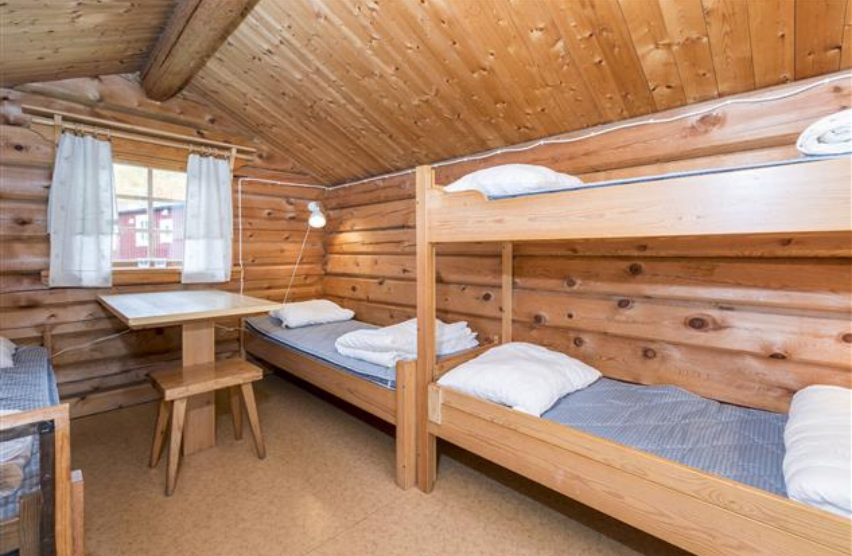 A wooden cabin room with bunk beds, a small table, and a window with curtains.