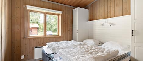 Une chambre avec des murs en bois, un grand lit avec une literie blanche, une fenêtre donnant sur une maison et une armoire blanche.