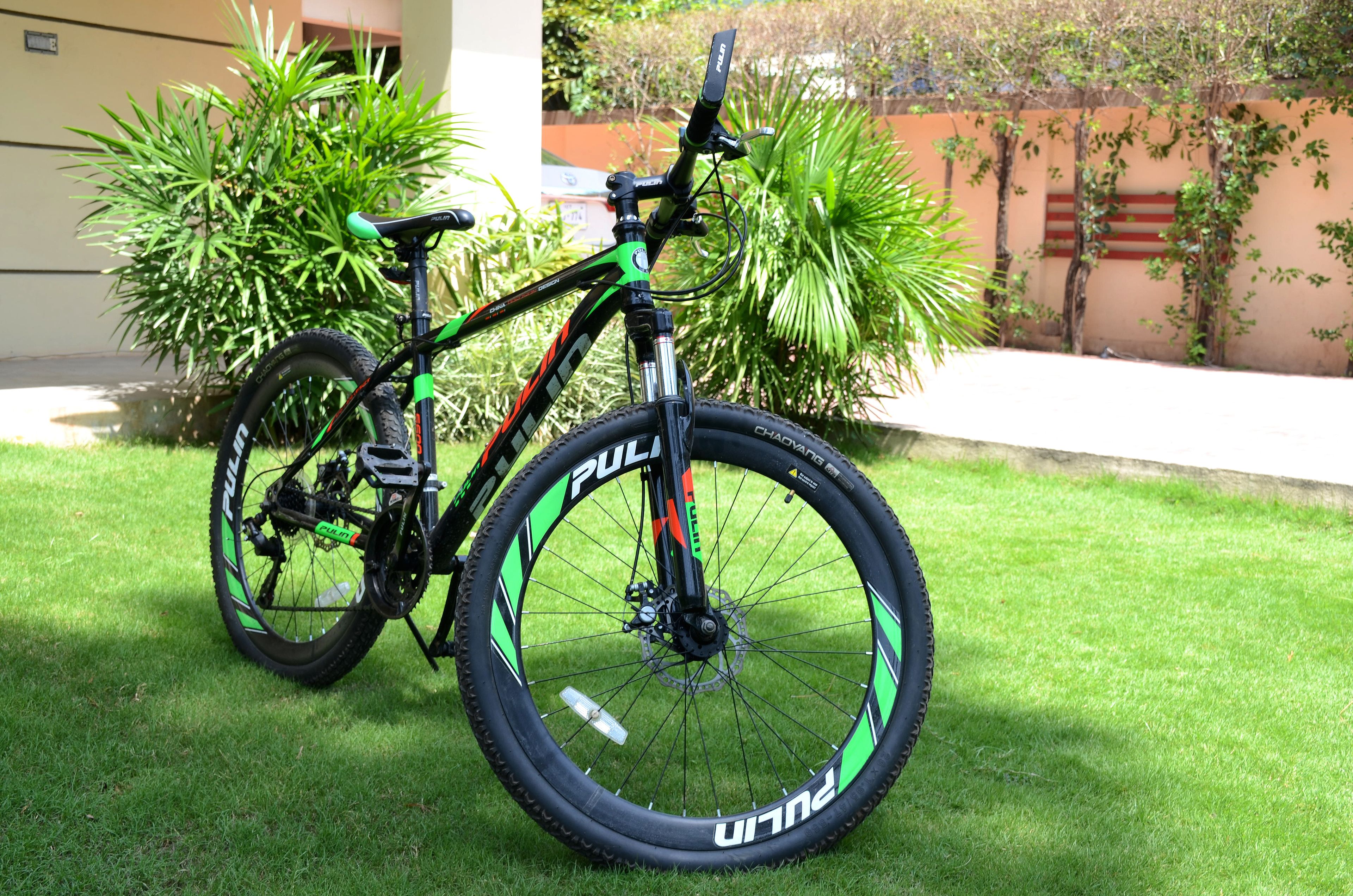 A bicycle with a green and black frame, black tires, and a front suspension fork, parked on a grassy lawn with a building and plants in the background.
