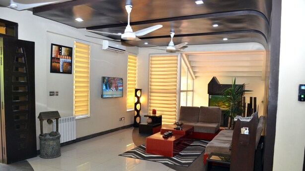 A modern living room with a sofa, coffee table, and decorative items. There are ceiling fans, a fireplace, and a large window with blinds. - Gofas Lodge (Islamabad)