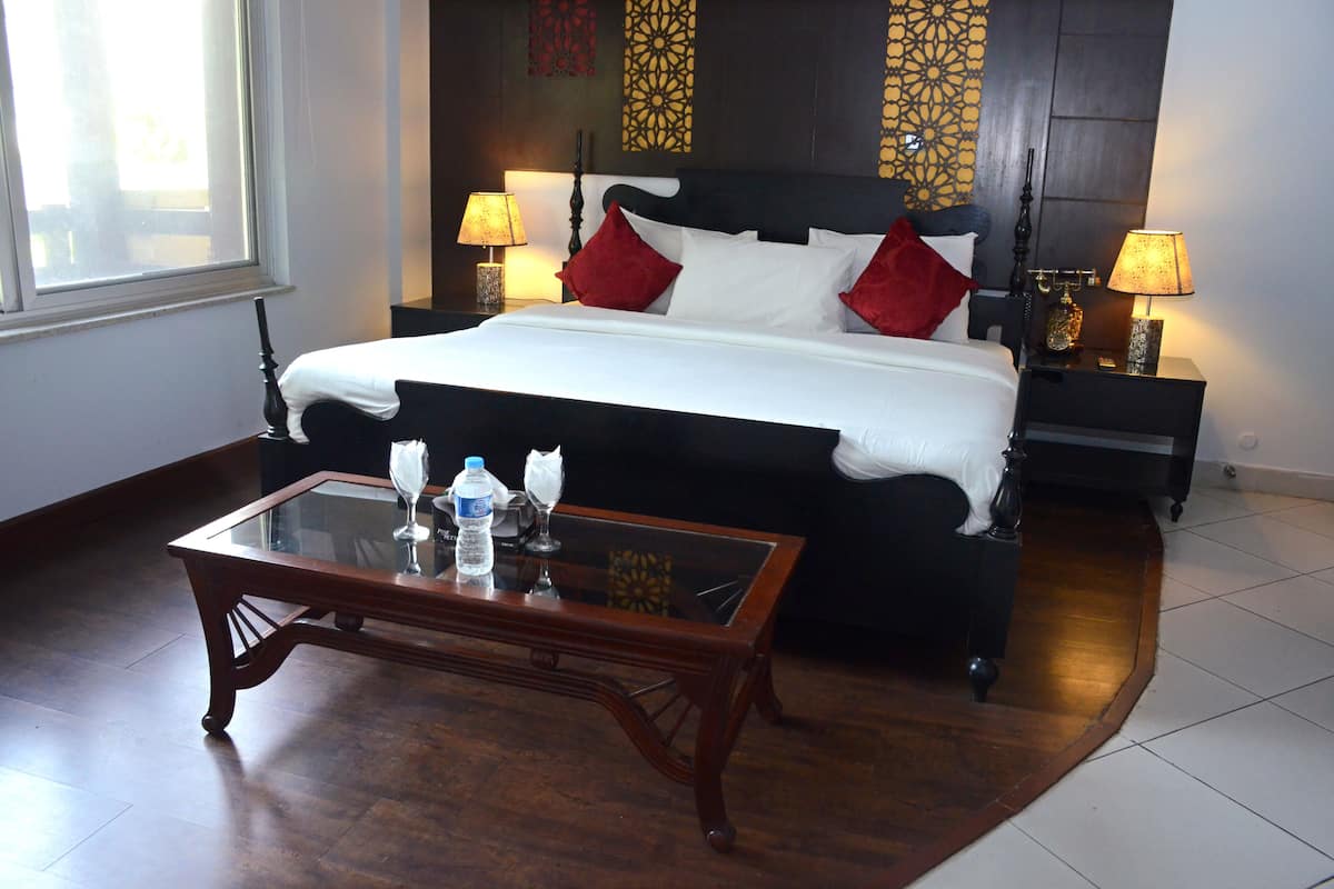 A hotel room with a large bed, two bedside tables with lamps, a glass coffee table, and a view of the outside through a window.