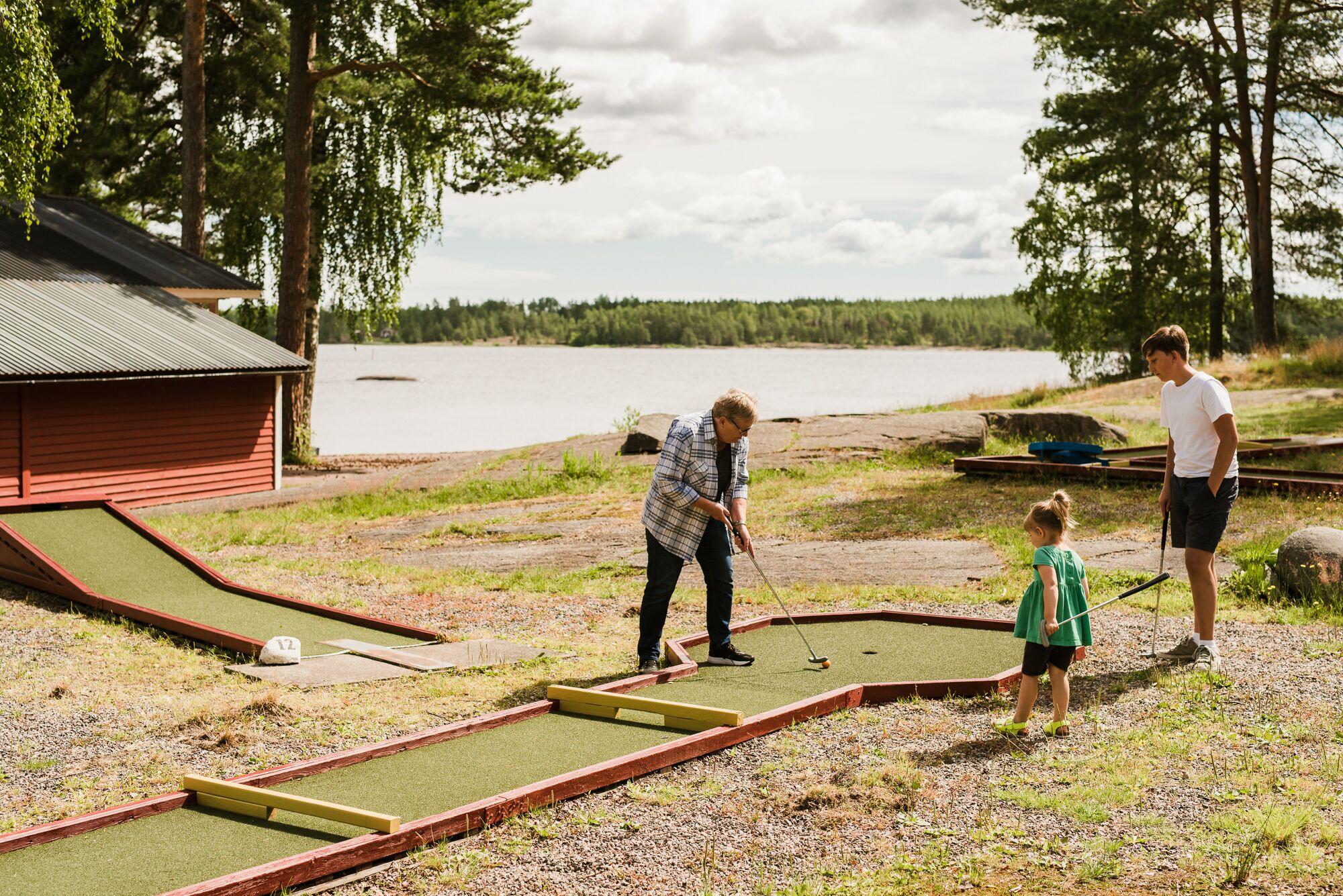 Un campo de mini golf con un edificio rojo, un estanque y árboles al fondo.