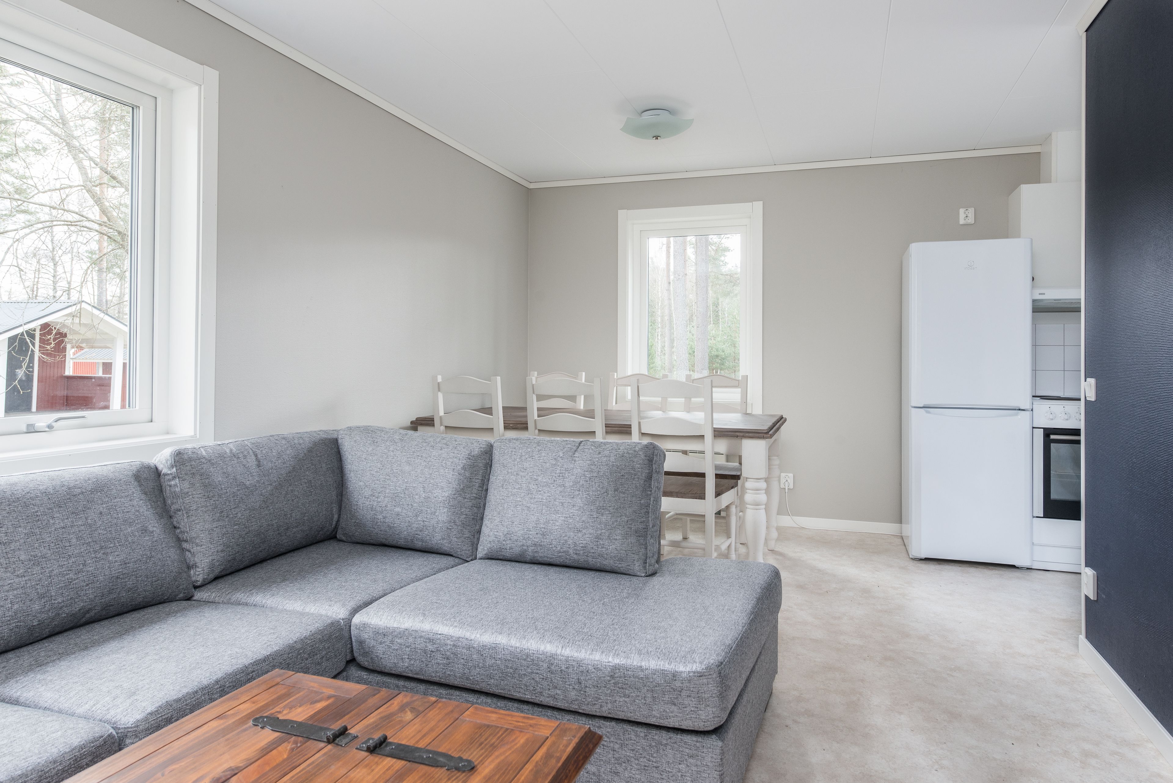 Una sala de estar moderna con un sofá seccional gris, una mesa de centro de madera, un comedor con sillas blancas y un refrigerador blanco.