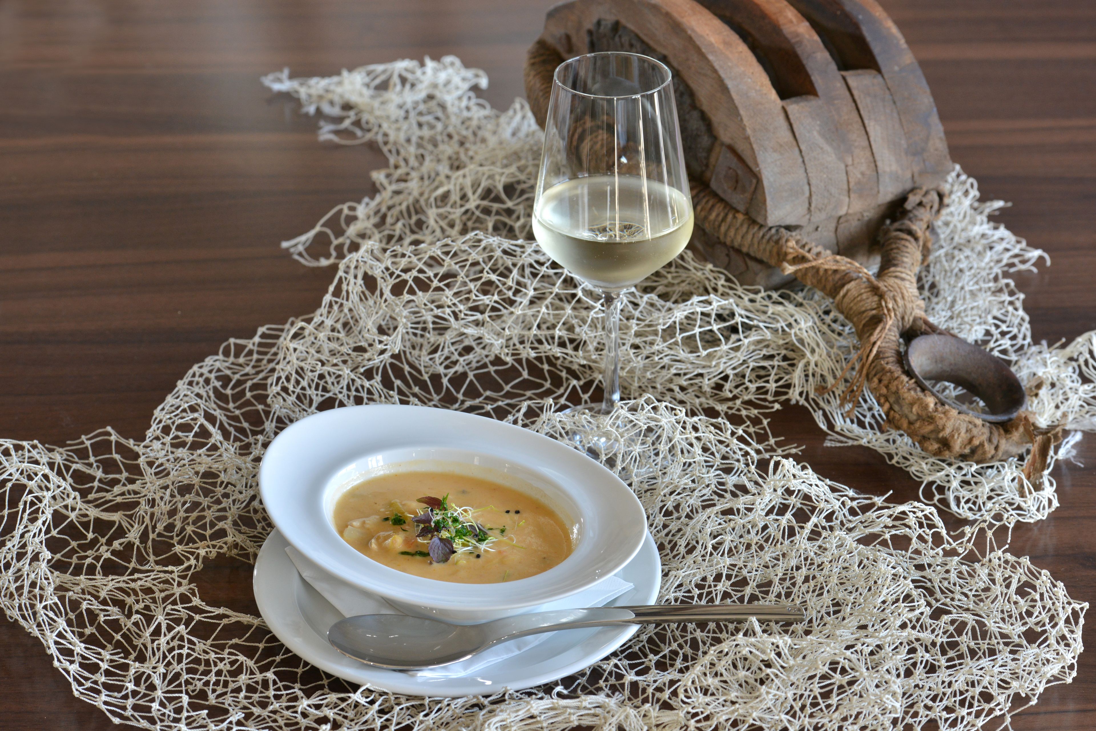 Un bol de soupe est posé sur une assiette blanche, accompagné d’une cuillère, le tout sur une table recouverte d’un tissu blanc à mailles. On aperçoit également un verre de vin blanc et un tonneau en bois.