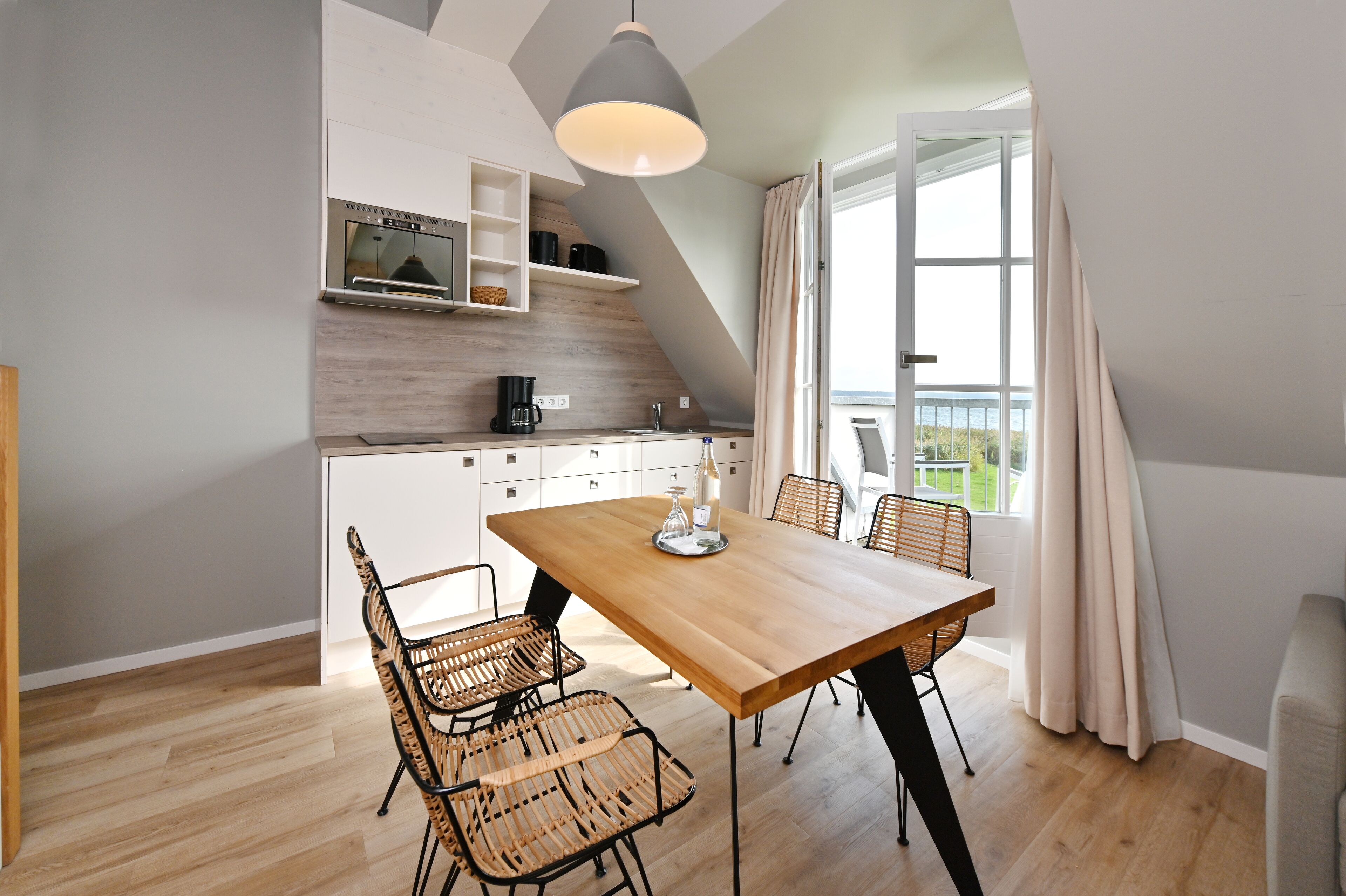 A modern kitchen with white cabinets, a wooden countertop, and a built-in oven. There is a dining area with a wooden table and wicker chairs.