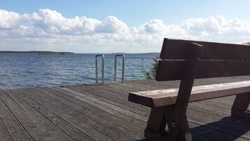 Une jetée en bois, avec un banc, des garde-corps en métal et une vue sur un lac paisible, avec une terre ferme visible au loin.