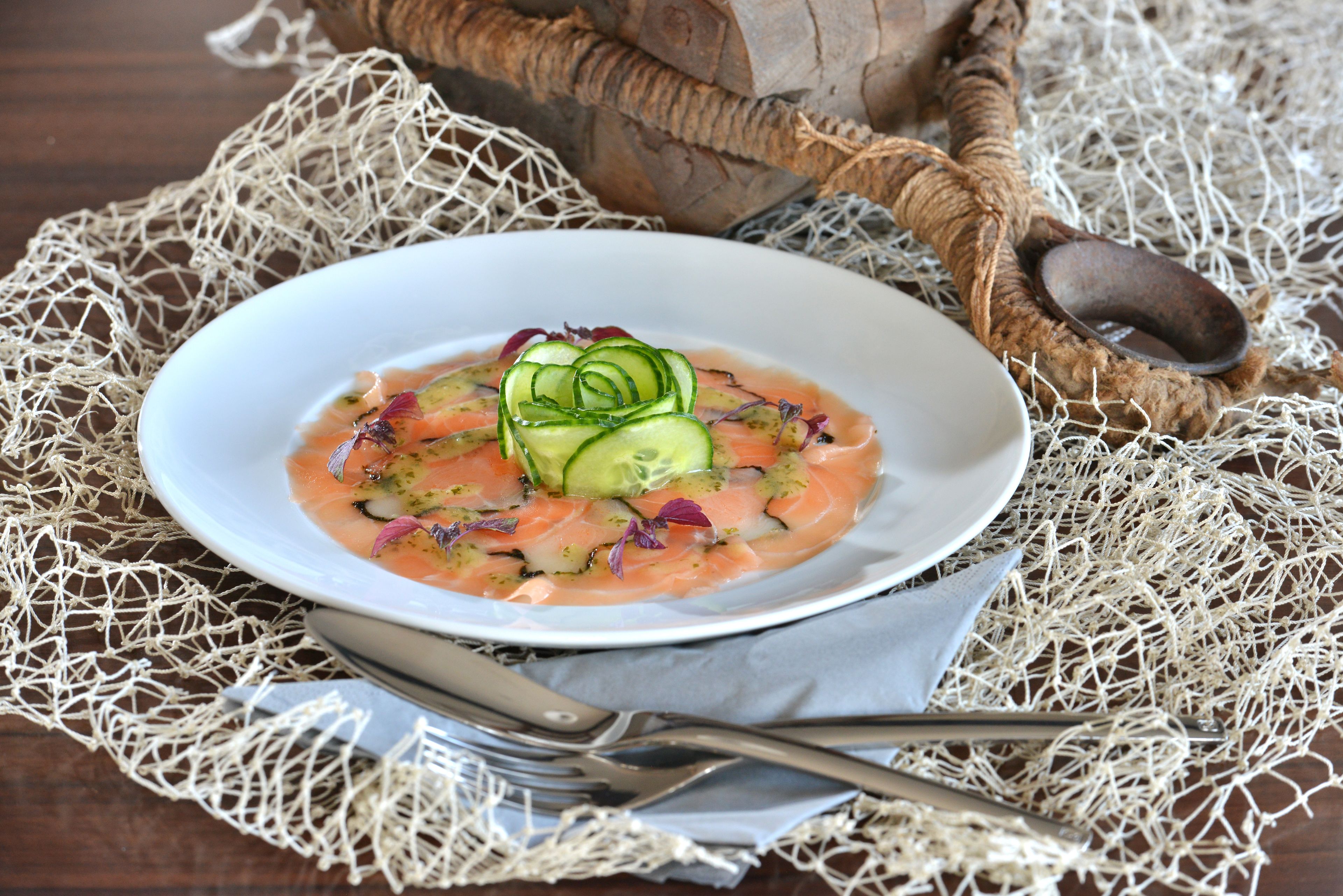 Un tazón blanco con tártara de salmón, pepino y cebolla roja sobre un mantel tejido, acompañado de un tenedor y una cuchara.